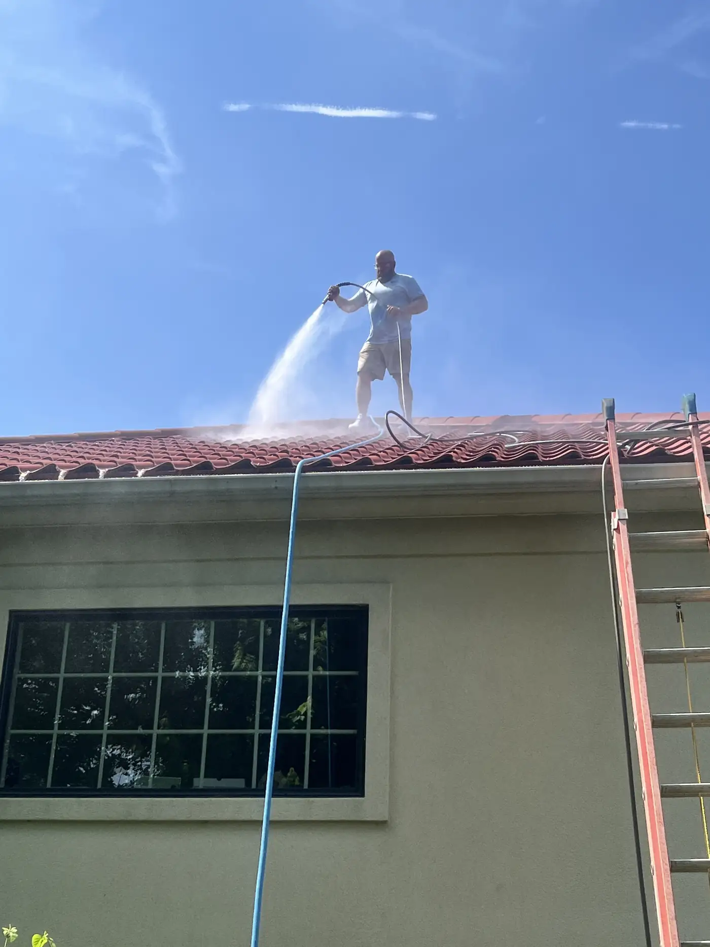 Crew member on a ladder washing a tall stucco wall under a tile roof edge