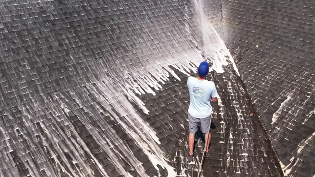 Crew member in a Revive shirt spraying cleaner on a dark, streaked shingle roof