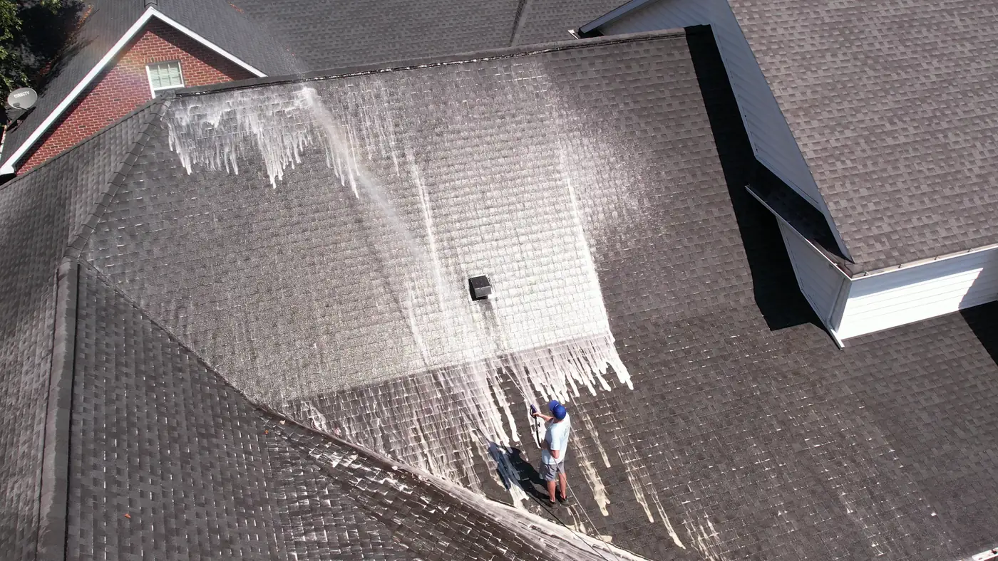 Drone view of a crew member soft washing a shingle roof, white cleaning foam running down the slope