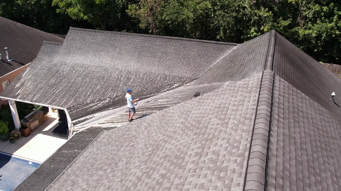 Drone view over a large shingle roof and backyard pool while a crew member soft washes one slope