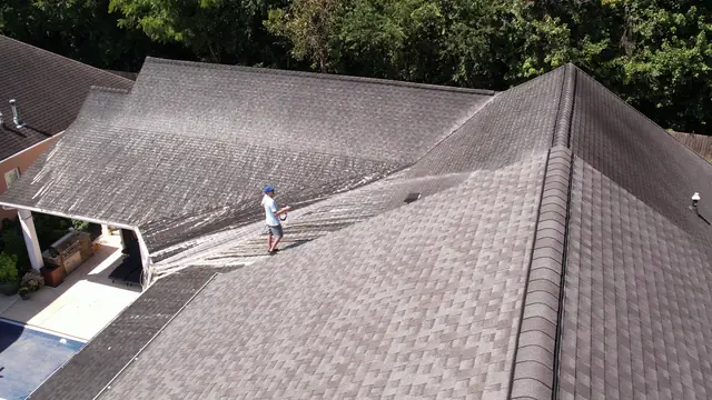 Drone view over a large shingle roof and backyard pool while a crew member soft washes one slope