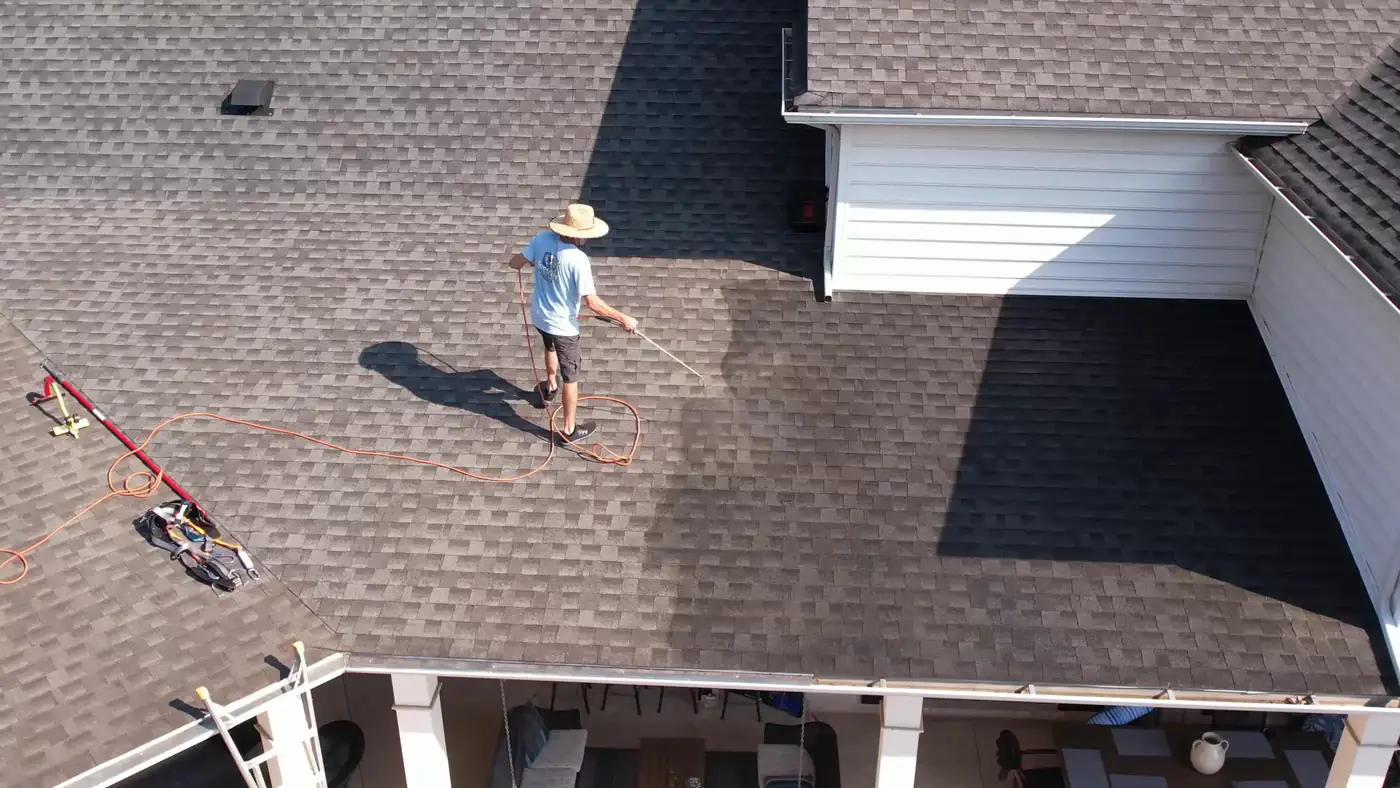 Crew member in a sun hat spraying treatment across a brown shingle roof, seen from above