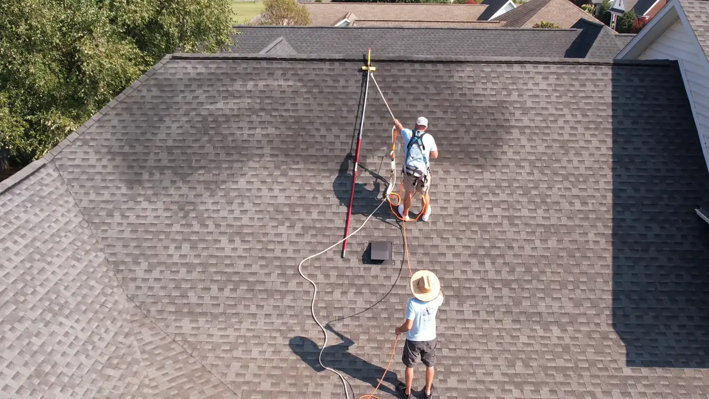 Drone view of two crew members applying a roof treatment with a sprayer and a pole on a shingle roof